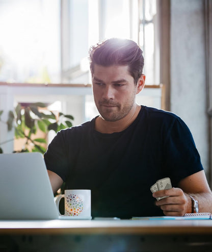 Man sitting at a desk with a laptop, holding money, in a bright room with large windows.